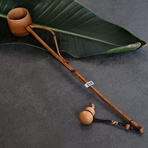 May include: A light brown bamboo water ladle with a long, slender handle and a deep, cylindrical cup. A small, light-colored wooden gourd-shaped charm hangs from the end of the handle by a black cord. A white sticker with the number 238 is attached.