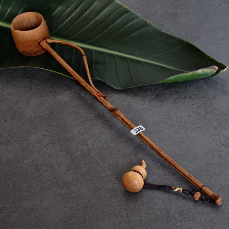 May include: A light brown bamboo water ladle with a long, slender handle and a deep, cylindrical cup. A small, light-colored wooden gourd-shaped charm hangs from the end of the handle by a black cord. A white sticker with the number 238 is attached.
