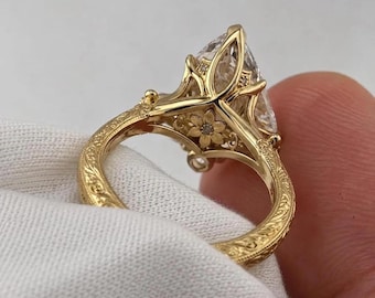 May include: A gold ring featuring a large pear-shaped center stone held by a decorative setting with floral motifs. The band and the basket of the ring are adorned with intricate engraved scrollwork patterns throughout the yellow gold metal surface.