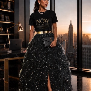 May include: A black t-shirt with the gold text THE NOW GENERATION, a metallic gold waist belt, and a voluminous black tulle maxi skirt featuring gold star-shaped embellishments. A black envelope-style clutch with a gold logo is held in front.
