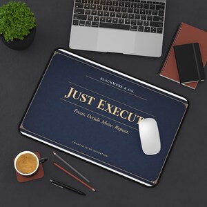 Könnte beinhalten: Ein rechteckiges, marineblaues Mauspad mit dem Text "JUST EXECUTE" in goldfarbenen Buchstaben. Das Mauspad trägt auch den Text "BLACKMERE & CO." und "Focus. Decide. Move. Repeat." Eine weiße Maus liegt auf dem Pad, mit einem Laptop und einem Notizblock im Hintergrund.