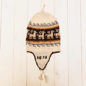 Könnte beinhalten: Eine beige Strickmütze mit Ohrenklappen und Quaste. Die Mütze hat ein braunes, beiges und graues geometrisches Muster mit Lamafiguren. Die Mütze hat eine spitze Oberseite und eine dunkelbraune Umrandung um die Ohrenklappen.
