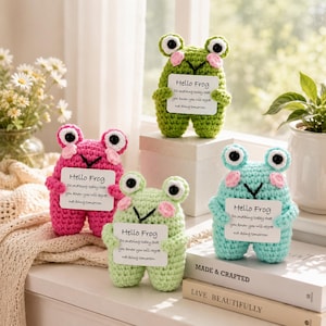 May include: Four crocheted frog figurines in green, pink, light green, and light blue. Each frog has large white eyes, pink cheeks, and holds a white rectangular sign that reads Hello Frog and Do something today that you know you will regret not doing tomorrow.