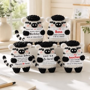 May include: Five crocheted zebra plush toys with black and white stripes, black manes, and black tails. Each zebra holds a white rectangular sign with black text, including phrases like You are ZE-best, Boss, and Have a wild Birthday.
