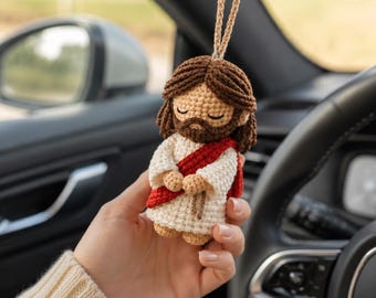 May include: A small, handmade crochet doll depicting a figure with brown hair and a beard, wearing a white robe with a red sash draped across the chest. The doll has closed eyes and is suspended by a tan string, designed as a hanging ornament.