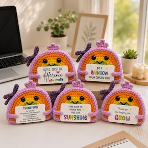 Op de afbeelding: Vijf halfronde gehaakte regenboogfiguren met paarse strikjes, zwarte ogen en lachende mondjes. Elk figuur houdt een wit rechthoekig kaartje vast met gedrukte tekst, waaronder zinnen zoals Never Forget The Difference You Make.