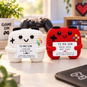 Op de afbeelding: Twee gehaakte knuffels in de vorm van gamecontrollers, één wit en één rood, elk met zwarte ogen en een glimlachende mond. Ze houden witte rechthoekige kaartjes vast met zwarte tekst "TO MY SON" en berichten over liefde voor gamen.