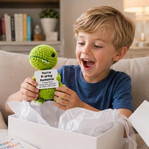 Op de afbeelding: Een groene gehaakte dinosauruspop met een wit label met de tekst "You're Rearing Awesome". De pop wordt vastgehouden door een kind dat lacht. De pop zit in een witte geschenkdoos met vloeipapier.