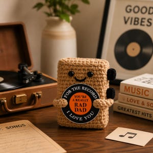 May include: A tan crocheted plush toy shaped like a record player, featuring black button eyes and a stitched smile. It holds a circular sign with black and orange text that reads: FOR THE RECORD YOU'RE A REALLY RAD DAD I LOVE YOU.