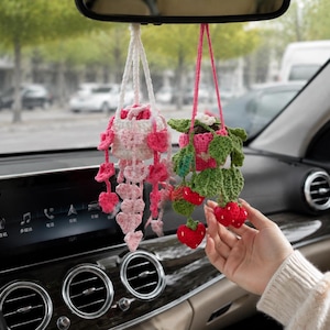 May include: Two hanging crochet plant ornaments for a car rearview mirror. One features a white pot with pink heart-shaped vines, while the other has a pink pot with green leaves and red strawberry accents, both suspended by yarn strings.