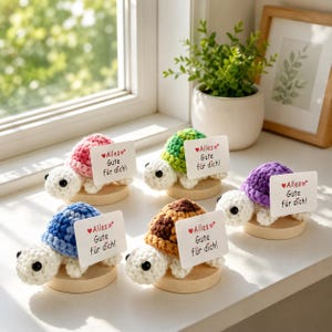 May include: A collection of handmade crochet turtle figurines in various colors, each holding a small card with German text. The turtles have white heads and feet, and sit on wooden bases. The text on the cards reads "Alles Gute für dich!"