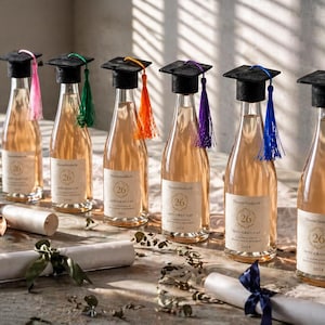 Peut inclure: Six bouteilles de vin avec des bouchons en forme de chapeaux de graduation de différentes couleurs. Chaque bouteille porte une étiquette avec le numéro 26. Des diplômes roulés avec des rubans sont au premier plan.