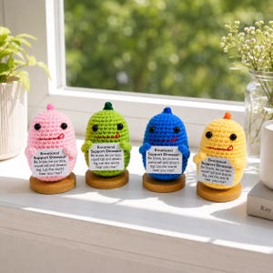 Op de afbeelding: Vier kleurrijke gehaakte dinosaurusfiguren in roze, groen, blauw en geel, elk met een bordje met de tekst "Emotional Support Dinosaur". Ze staan op houten voetstukken.