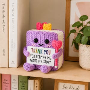 May include: A purple crocheted book-shaped plush toy with black eyes, a smiling mouth, and pink cheeks. It features a yellow and pink bookmark on top, holds a pink heart-shaped wand, and displays a white sign reading THANK YOU FOR HELPING ME WRITE MY STORY.