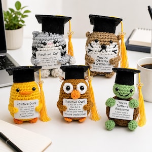 Può includere: Una collezione di cinque figurine di animali all'uncinetto a tema laurea. Ogni figura, tra cui un gatto, una tigre, un'anatra, un gufo e una tartaruga, indossa un cappello di laurea nero e tiene in mano un cartello con un messaggio motivazionale.