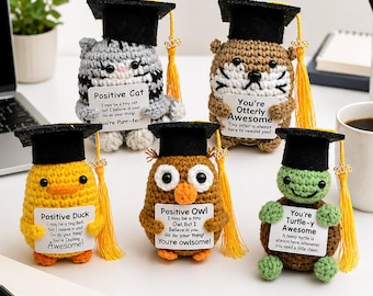Può includere: Una collezione di cinque figurine di animali all'uncinetto a tema laurea. Ogni figura, tra cui un gatto, una tigre, un'anatra, un gufo e una tartaruga, indossa un cappello di laurea nero e tiene in mano un cartello con un messaggio motivazionale.