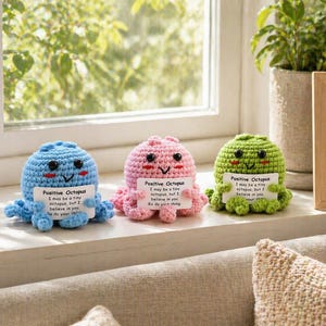 May include: Three crocheted octopus figurines in blue, pink, and green, each holding a small sign with the text "Positive Octopus". The octopus have smiling faces and are displayed on a windowsill with a plant and a framed picture in the background.
