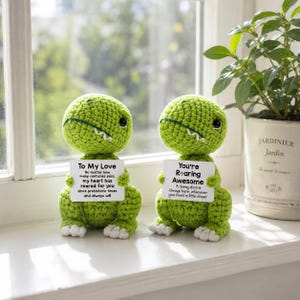 May include: Two green crocheted dinosaur figures with white teeth and eyes. Each dinosaur holds a sign with text. One says "To My Love" and the other says "You're Roaring Awesome". The dinosaurs are on a windowsill.