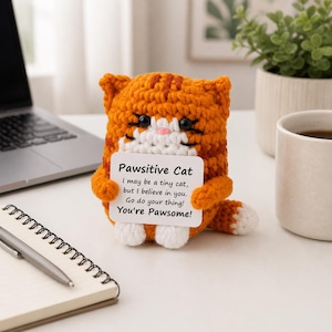 Può includere: Figurina di gatto tabby arancione all'uncinetto che tiene in mano un biglietto bianco con la scritta "Pawsitive Cat". Il gatto ha occhi neri, naso rosa e zampe bianche. Il biglietto recita "I may be a tiny cat, but I believe in you. Go do your thing! You're Pawsome!"