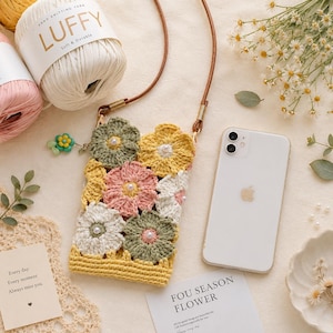 Op de afbeelding: Een rechthoekig gehaakt telefoonhoesje met een bloemenpatroon van saliegroene, gele en zachtroze garenbloemen, elk in het midden voorzien van een kleine witte parel. Het hoesje heeft een dunne bruine leren riem en goudkleurige metalen bevestigingen.