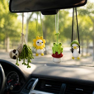 May include: Four crocheted hanging ornaments suspended from a rearview mirror. The set includes a sloth in a green plant hanger, a white creature with a yellow flower head, a green frog sitting on a red mushroom, and a white duck on a black swing.