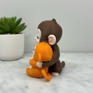 Pode incluir: Uma estatueta de macaco marrom abra&ccedil;ando uma estatueta de macaco laranja. As estatuetas est&atilde;o em uma superf&iacute;cie branca com uma pequena planta suculenta em vaso ao fundo. Os macacos t&ecirc;m detalhes faciais brancos.