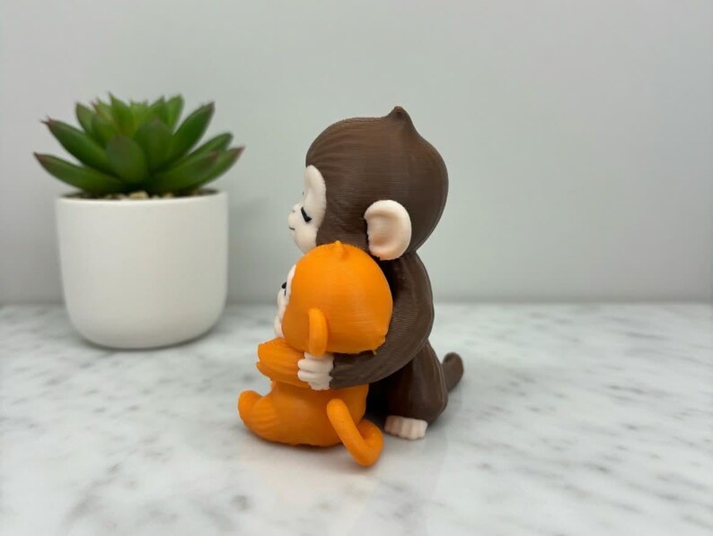 Pode incluir: Uma estatueta de macaco marrom abra&ccedil;ando uma estatueta de macaco laranja. As estatuetas est&atilde;o em uma superf&iacute;cie branca com uma pequena planta suculenta em vaso ao fundo. Os macacos t&ecirc;m detalhes faciais brancos.