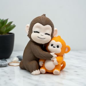 Pode incluir: Uma estatueta de macaco marrom e laranja abra&ccedil;ando outra estatueta de macaco laranja. As estatuetas t&ecirc;m rostos brancos e est&atilde;o sorrindo. O fundo inclui uma pequena planta em vaso, seixos e um anel de ouro.