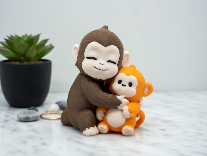 Pode incluir: Uma estatueta de macaco marrom e laranja abra&ccedil;ando outra estatueta de macaco laranja. As estatuetas t&ecirc;m rostos brancos e est&atilde;o sorrindo. O fundo inclui uma pequena planta em vaso, seixos e um anel de ouro.