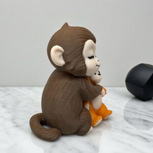 Pode incluir: Uma estatueta de macaco marrom abra&ccedil;ando uma estatueta de macaco laranja. O macaco marrom tem orelhas brancas e um rosto sorridente. Um objeto geom&eacute;trico preto est&aacute; &agrave; direita dos macacos.