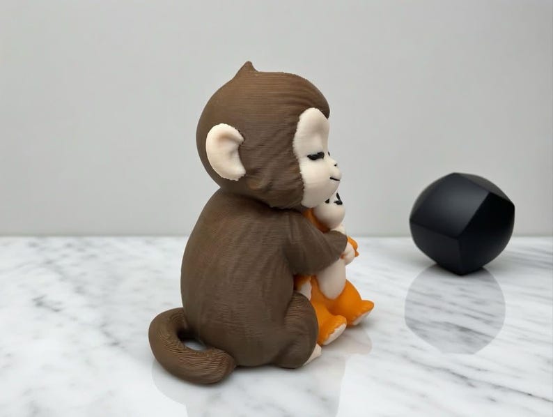 Pode incluir: Uma estatueta de macaco marrom abra&ccedil;ando uma estatueta de macaco laranja. O macaco marrom tem orelhas brancas e um rosto sorridente. Um objeto geom&eacute;trico preto est&aacute; &agrave; direita dos macacos.