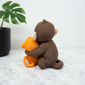 Pode incluir: Uma estatueta de macaco marrom abra&ccedil;ando uma estatueta de macaco laranja. O macaco marrom tem orelhas brancas e uma cauda pequena. O macaco laranja tem uma cauda curva e est&aacute; sendo abra&ccedil;ado pelo macaco marrom. Uma planta em vaso com folhas verdes est&aacute; no fundo.