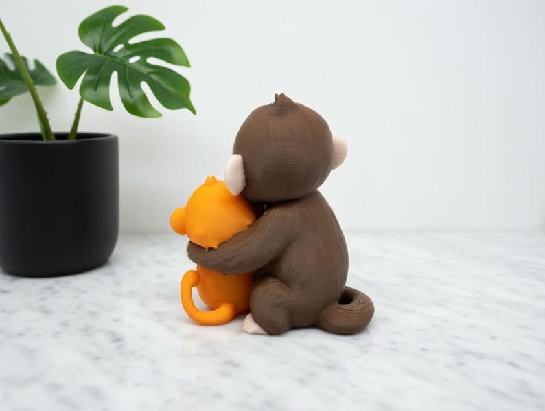 Pode incluir: Uma estatueta de macaco marrom abra&ccedil;ando uma estatueta de macaco laranja. O macaco marrom tem orelhas brancas e uma cauda pequena. O macaco laranja tem uma cauda curva e est&aacute; sendo abra&ccedil;ado pelo macaco marrom. Uma planta em vaso com folhas verdes est&aacute; no fundo.