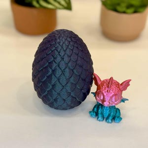 May include: A dark blue, textured dragon egg next to a small, colorful dragon figurine. The dragon is pink and blue with large eyes and small wings. The egg is oval-shaped and has a scale-like pattern.
