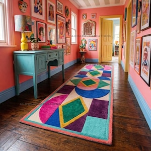 May include: A colorful geometric patterned rug with a cream background, featuring shapes in pink, green, blue, purple, and yellow. The rug is rectangular and placed on a dark wood floor in a hallway. The walls are painted coral and adorned with framed artwork.