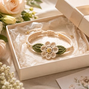 Könnte beinhalten: Ein cremefarbenes Häkelarmband mit einem zentralen Blumenmotiv, das in der Mitte eine goldfarbene Perle aufweist und von zwei dunkelgrünen gehäkelten Blättern flankiert wird. Das verstellbare Band enthält kleine goldfarbene Perlen.