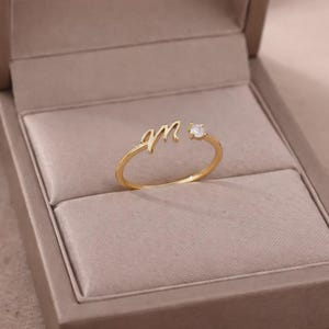 May include: A gold ring with the cursive letter "M" and a small, clear gemstone. The ring is presented in a beige jewellery box. The ring is a delicate piece of jewellery.