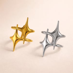 May include: Two rings, one gold-toned and one silver-toned, each featuring a double starburst design. The starbursts have pointed tips and are connected by a band that wraps around the finger. The rings are displayed against a neutral background.