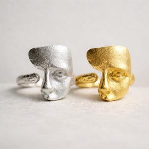 May include: Two rings, one silver and one gold, each with a sculpted face design. The faces have a minimalist, artistic style. The rings are set against a neutral background, highlighting the jewellery.