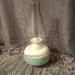 May include: A ceramic oil lamp featuring a rounded base with a white top and a turquoise and brown glazed bottom. The lamp includes a clear glass chimney that sits atop a white ceramic burner assembly with a small wick visible in the center.