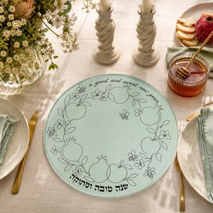 May include: A round, light green glass serving platter featuring a black line art illustration of pomegranates, apples, flowers, and bees. The text reads "a good and sweet new year" in English and "שנה טובה ומתוקה" in Hebrew script.