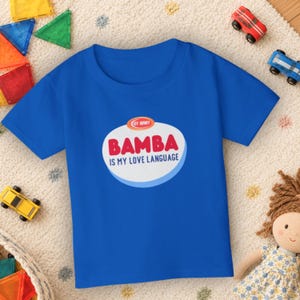 May include: A bright blue short-sleeved t-shirt featuring a central white oval graphic with a red orange logo at the top. The text reads BAMBA IS MY LOVE LANGUAGE in bold red and dark blue lettering, centered on the chest of the garment.