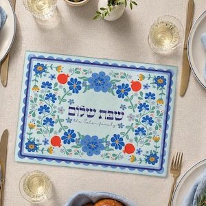 May include: A rectangular placemat with a light blue background, adorned with a floral design in blue, orange, and yellow. The placemat features the Hebrew text "Shabbat Shalom" and the family name "the Cohen family."