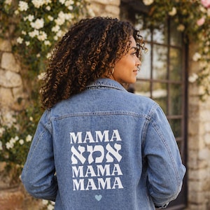 May include: A light blue denim jacket with the word "MAMA" printed in white in both English and Hebrew. The jacket has a small blue heart at the bottom. The jacket is a classic style with a button-down front and pockets.
