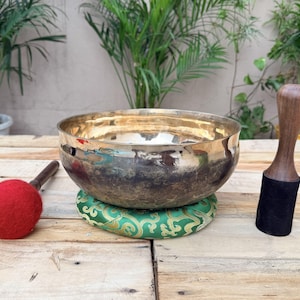 May include: A metallic singing bowl with a polished gold interior and a dark, weathered exterior finish. The bowl rests on a green patterned fabric cushion, accompanied by a red felt-tipped mallet and a wooden mallet with a black base.
