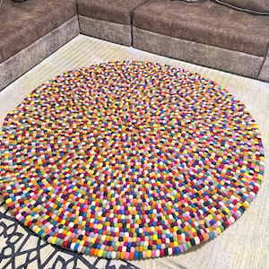 Op de afbeelding: Een ronde, kleurrijke vloerkleed met een dicht patroon van kleine, dicht opeengepakte viltballen. Het vloerkleed heeft een levendige mix van kleuren, waaronder rood, geel, blauw, groen en paars. Het vloerkleed ligt op een lichtgekleurde houten vloer.