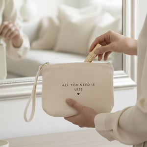 Könnte beinhalten: Eine beigefarbene Kosmetiktasche aus Canvas mit goldenem Reißverschluss und Handschlaufe. Auf der Tasche steht der Text "ALL YOU NEED IS LESS" und ein kleines Herz. Ein goldener Lippenstift wird in die Tasche gelegt.
