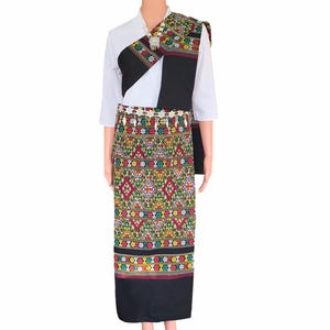 Puede incluir: Un atuendo tradicional tailandés exhibido en un maniquí. El conjunto incluye una blusa blanca, un chal negro y estampado, y una falda larga con un diseño geométrico vibrante en rojo, verde, amarillo y negro. Un cinturón decorativo añade detalles.