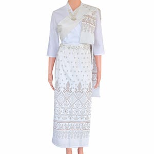 May include: White traditional Thai outfit, including a long skirt, top, and shawl. The skirt and shawl feature a detailed brown and white pattern. The outfit is accessorized with a silver belt and necklace. The top has three-quarter sleeves.