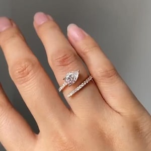 以下が含まれることがあります： ペアシェイプのダイヤモンドと小さなダイヤモンドのバンドが付いたローズゴールドのリング。指にはめられ、デザインと輝きを際立たせています。バンドは指に巻き付き、ユニークでエレガントな外観を作り出しています。